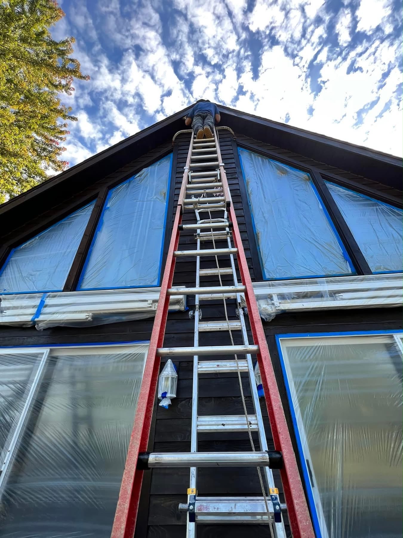 Crew member on an extension ladder staining a two-story gable, windows masked for protection