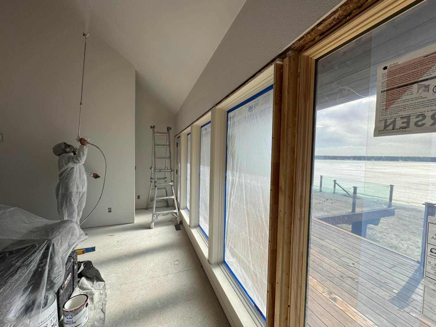 Sunroom interior painting in progress with drop cloths, ladders, and windows overlooking a frozen lake